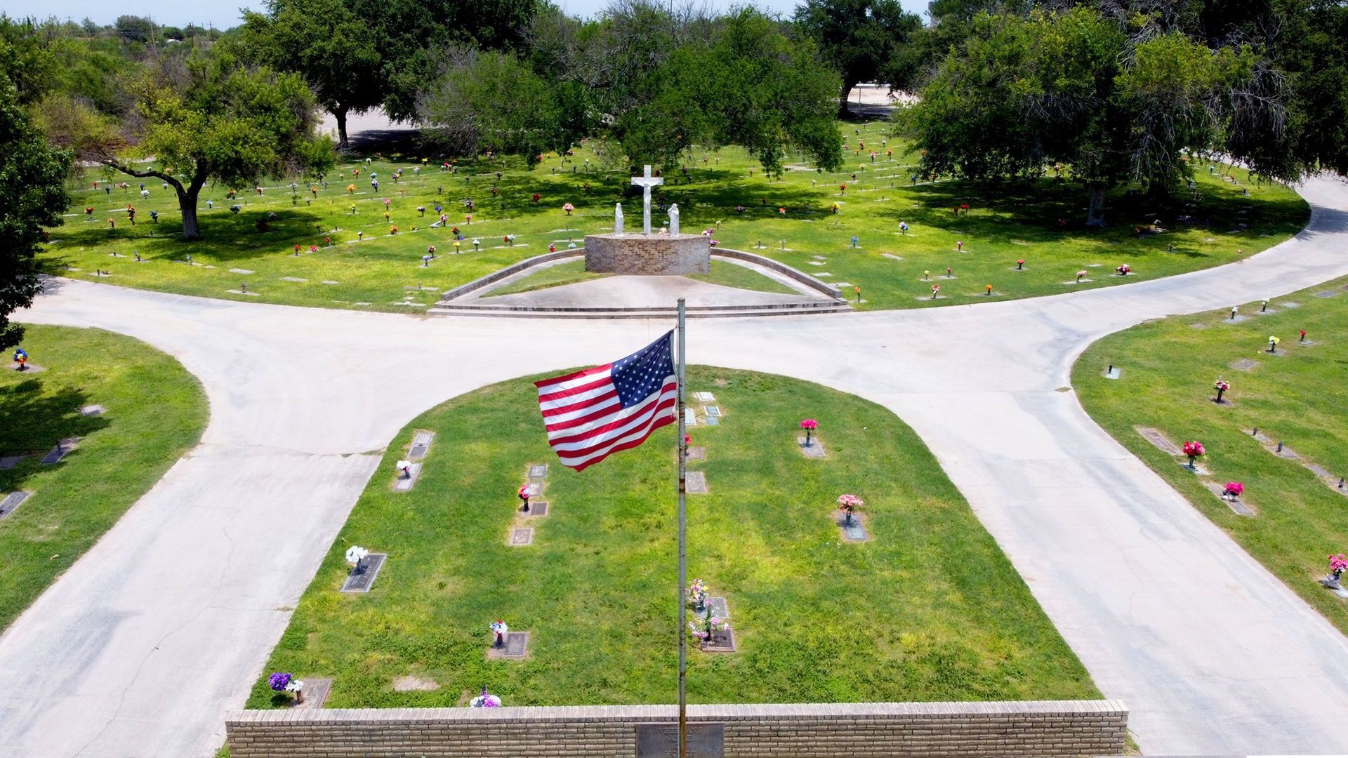 American flag flying at a cemetery entrance, with white roads and green trees in the background