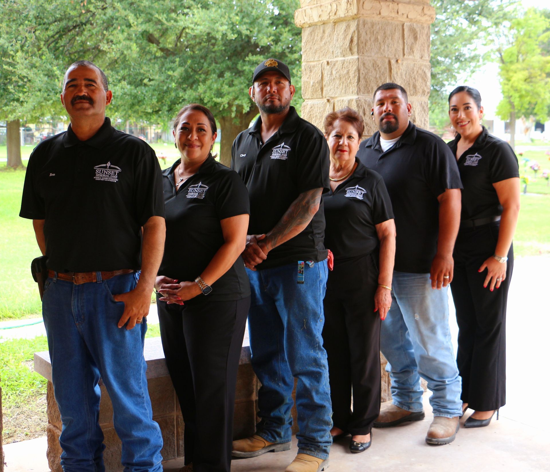 Six people in black shirts stand in a line under a stone pavilion, smiling outdoors.