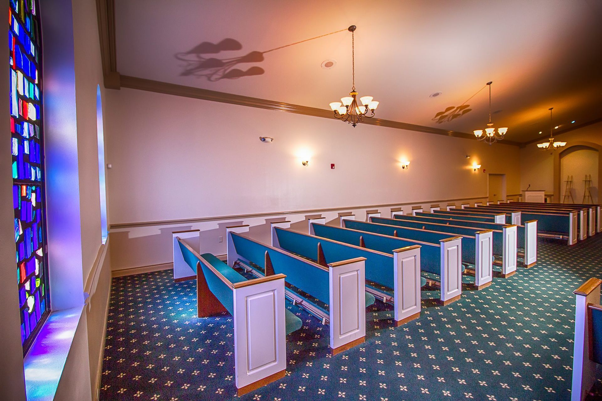 Empty church sanctuary with blue pews, stained-glass windows, and hanging lights