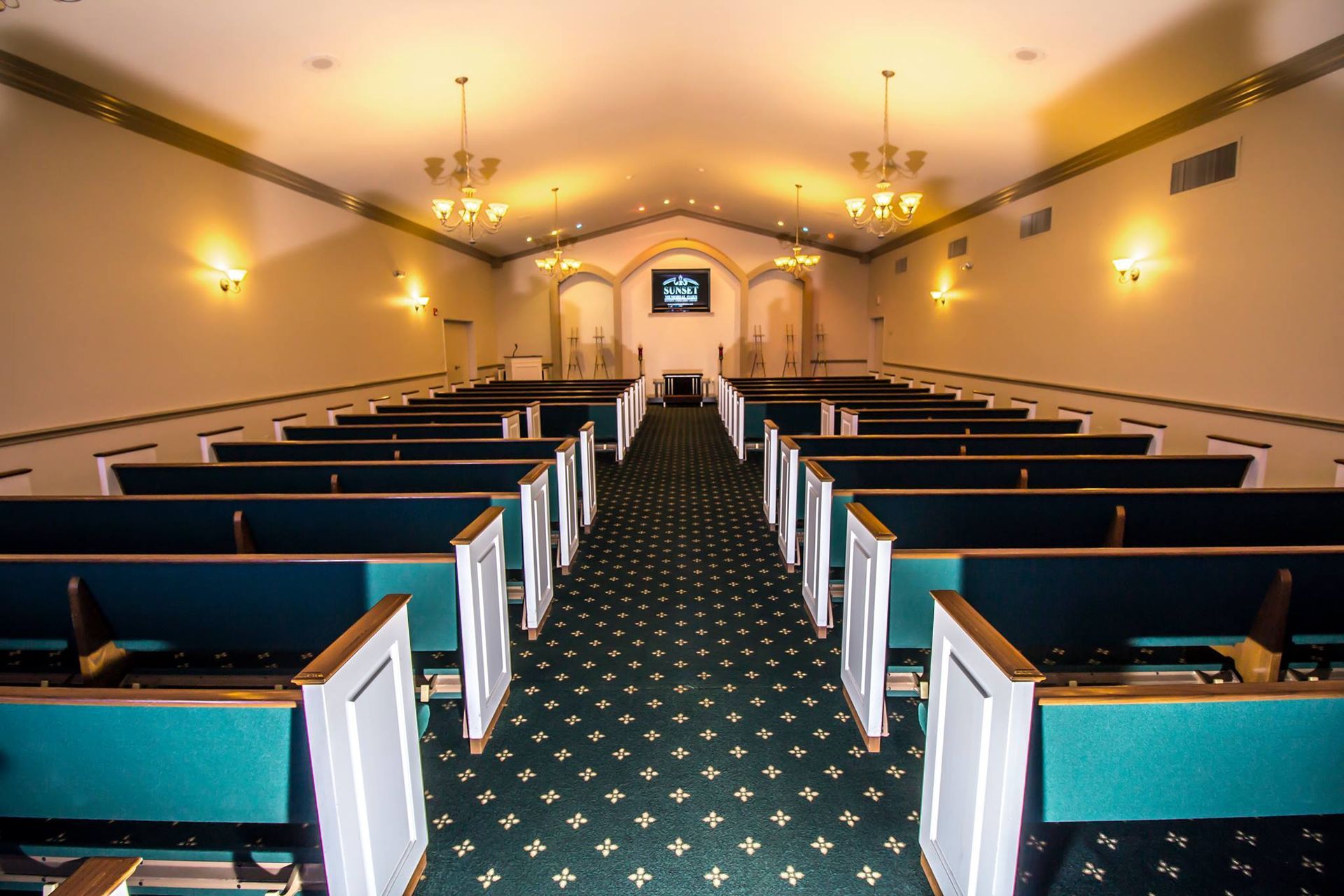 Empty church sanctuary with wooden pews, chandeliers, and a patterned green aisle carpet