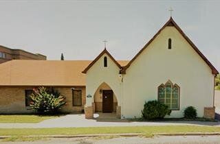 Small beige church with a steep roof and arched entrance beside a low brick building under a clear sky.