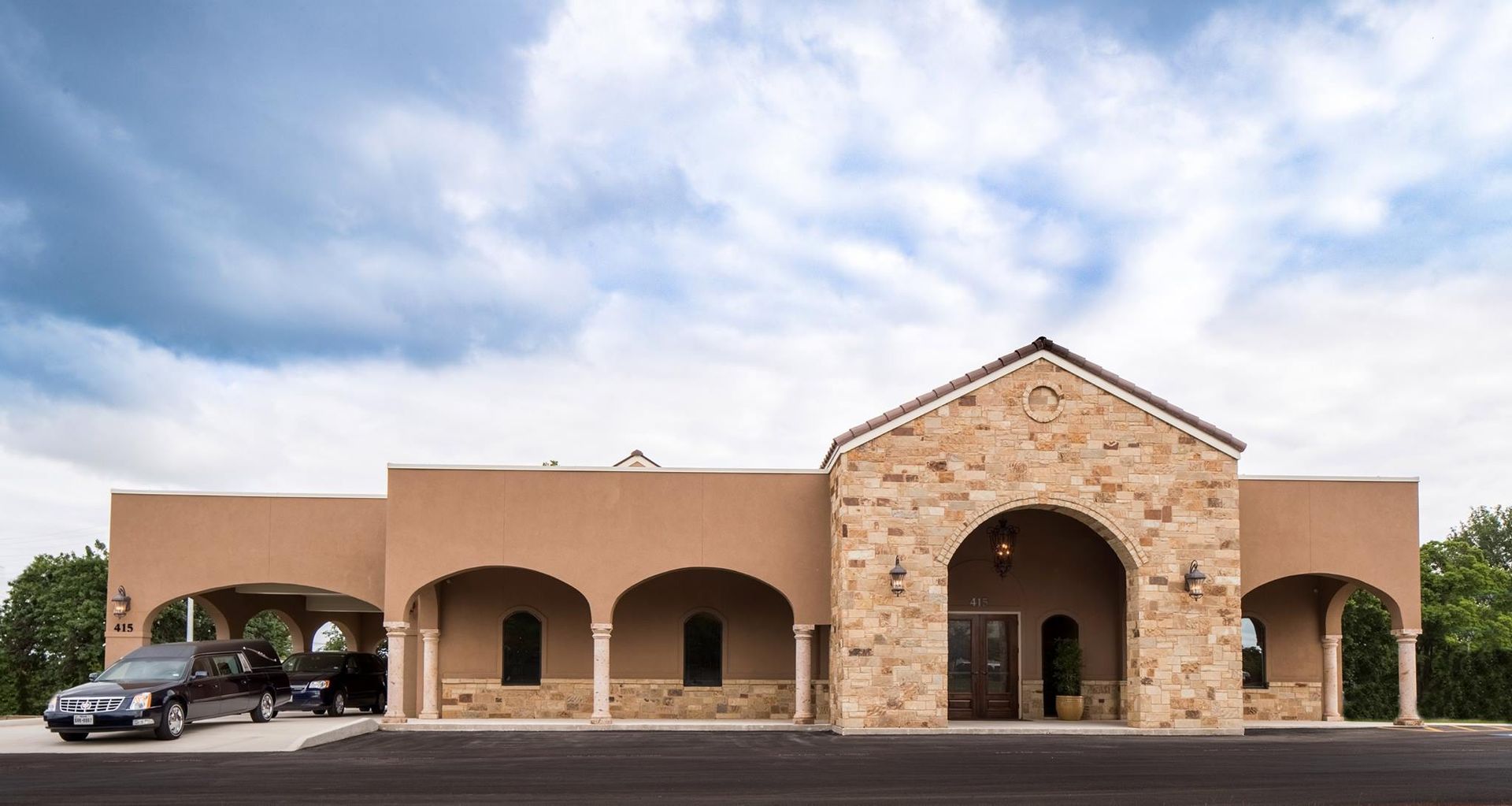 Stone-front building with arched entrances and a parked car under a cloudy sky