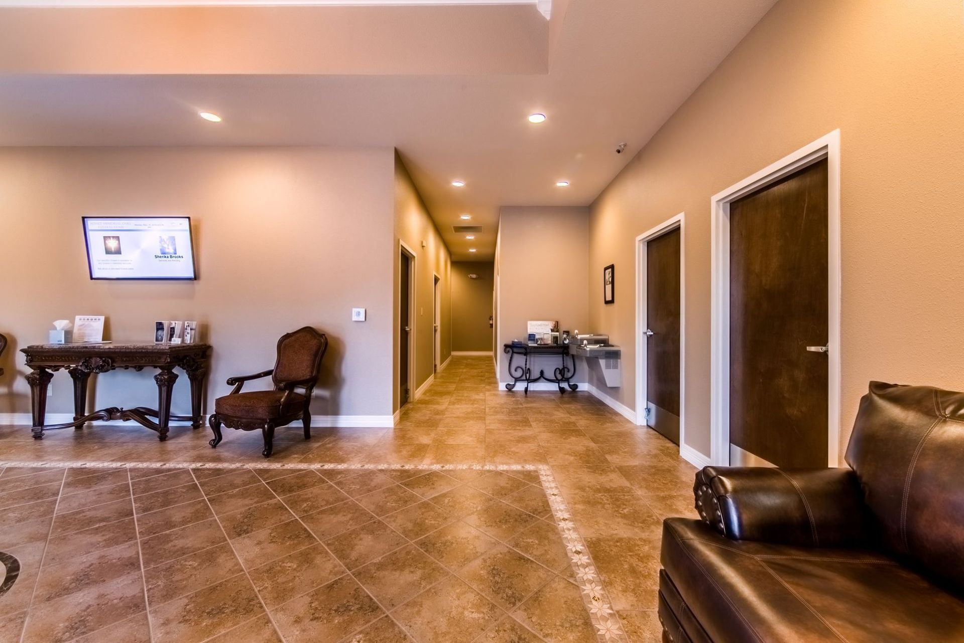 Spacious beige living room with tiled floor, dark furniture, hallway, and double doors.