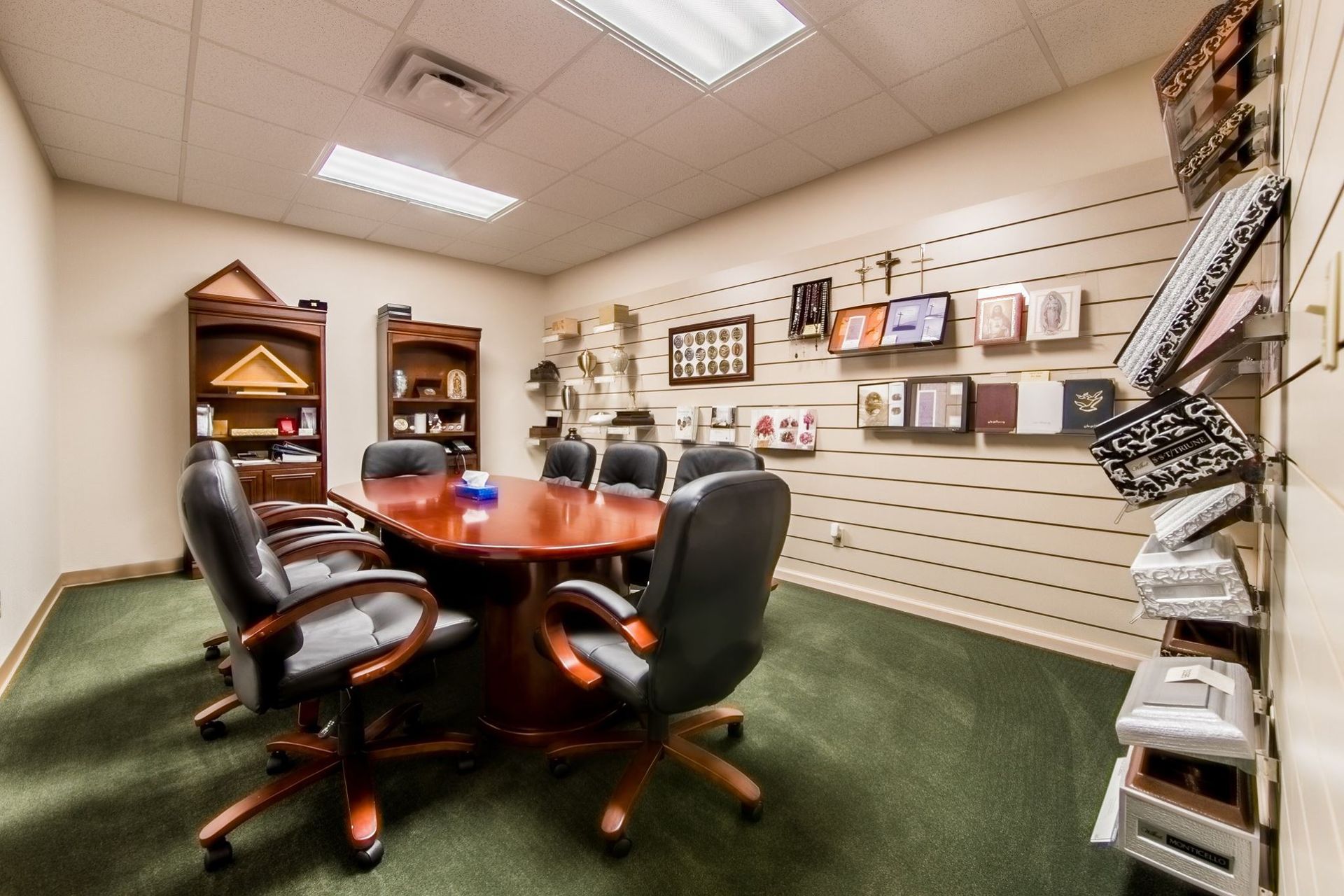 Small office conference room with round wooden table, black chairs, green carpet, and wall-mounted product samples.