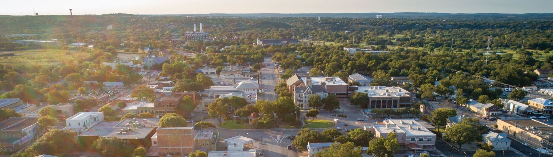 Aerial view of a suburban neighborhood at sunset, with trees, rooftops, and distant horizon.