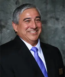 Smiling man in a dark suit with a blue tie against a gray studio backdrop