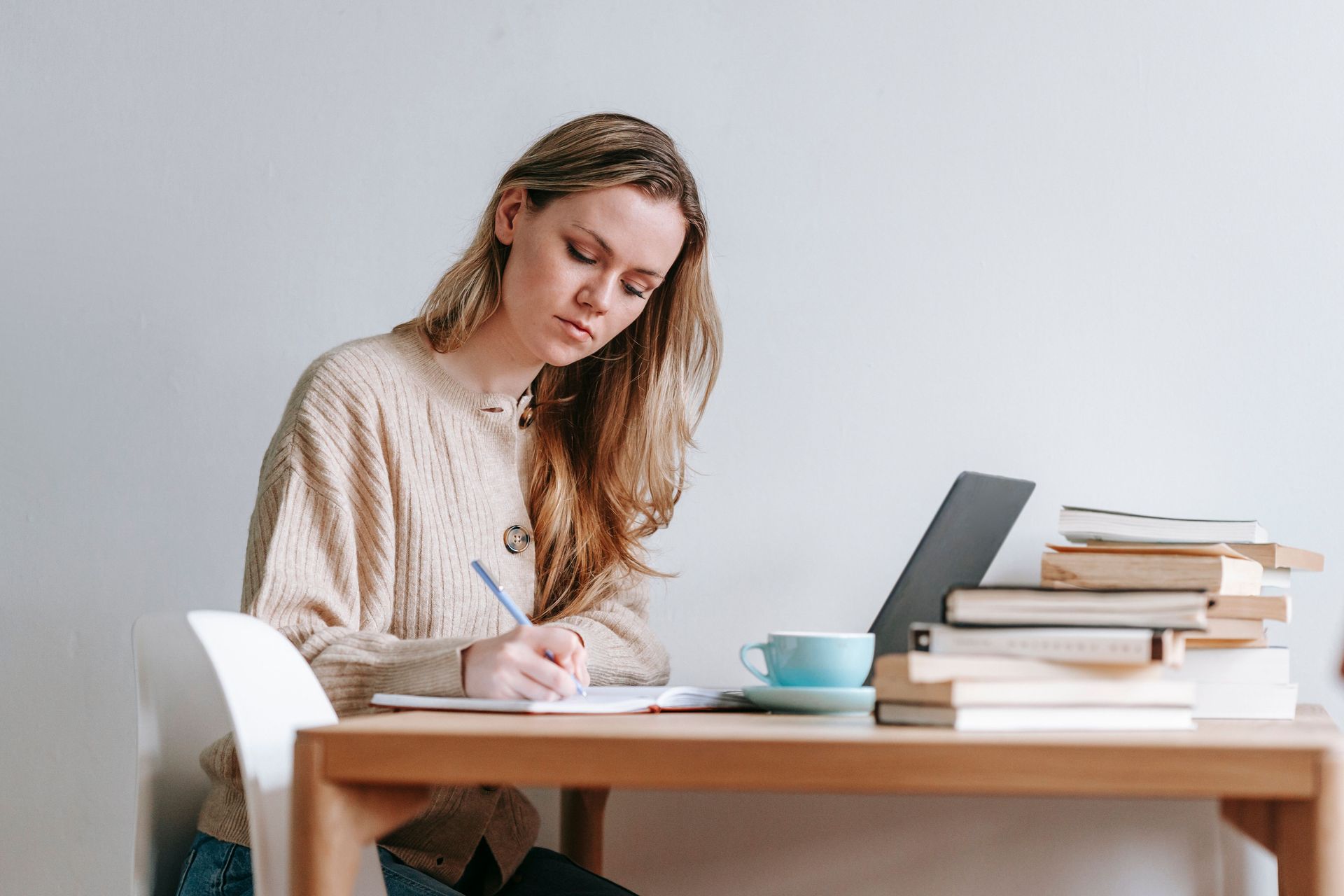 Woman writing at a wooden desk with a laptop, stack of books, and a cup in a bright room