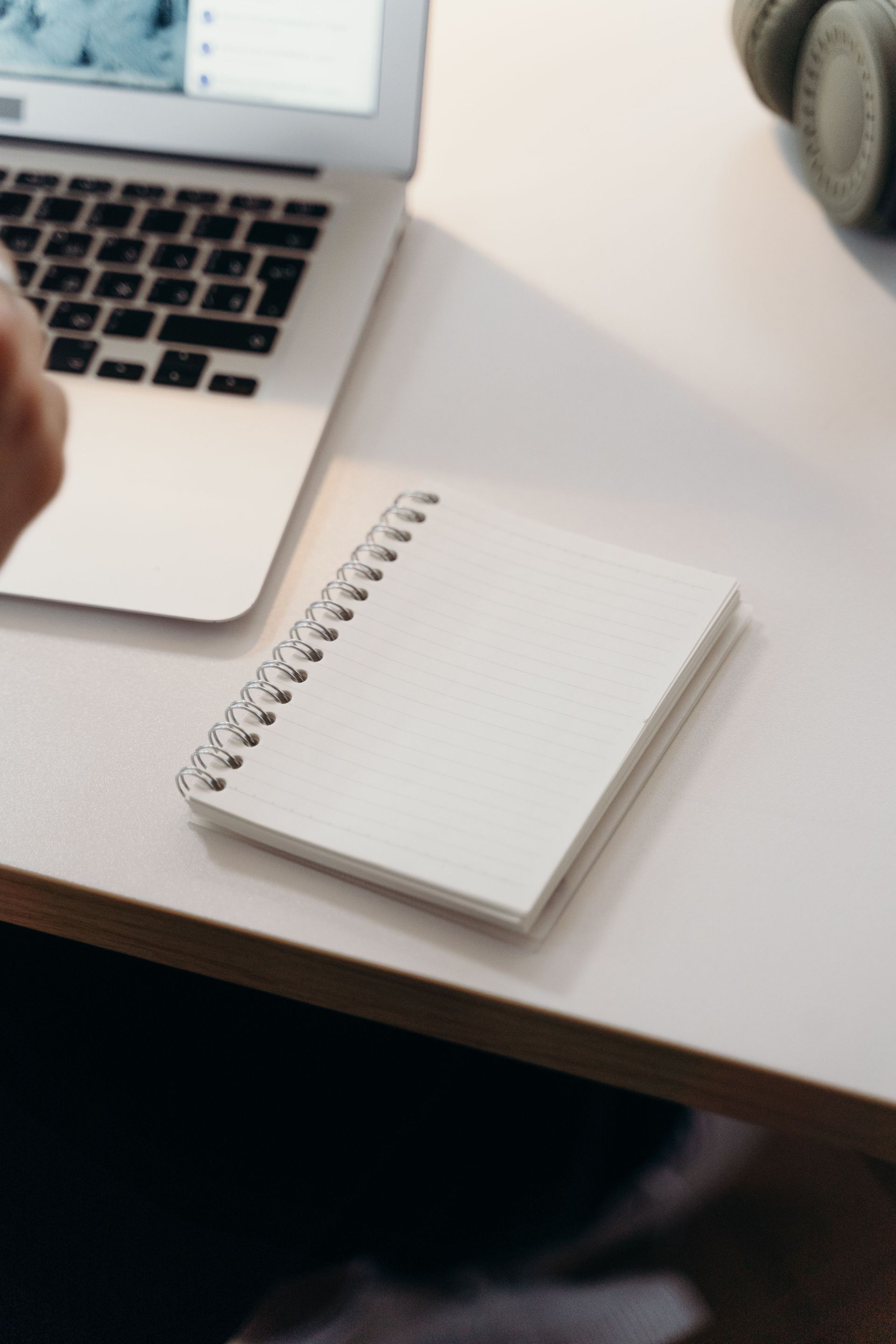 Laptop beside a blank spiral notebook on a white desk with headphones nearby