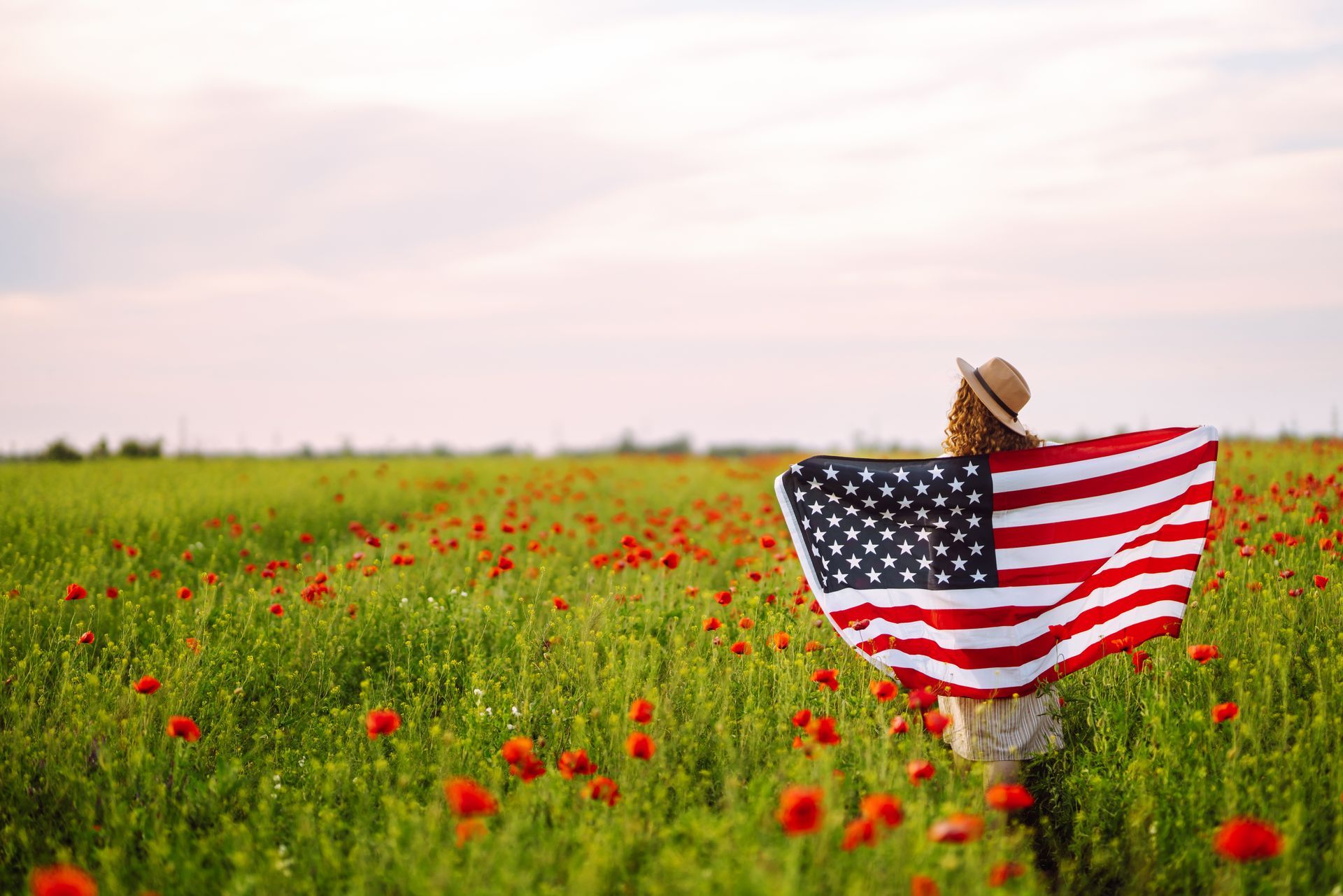 Person holding an American flag in a green field with red wildflowers at sunset