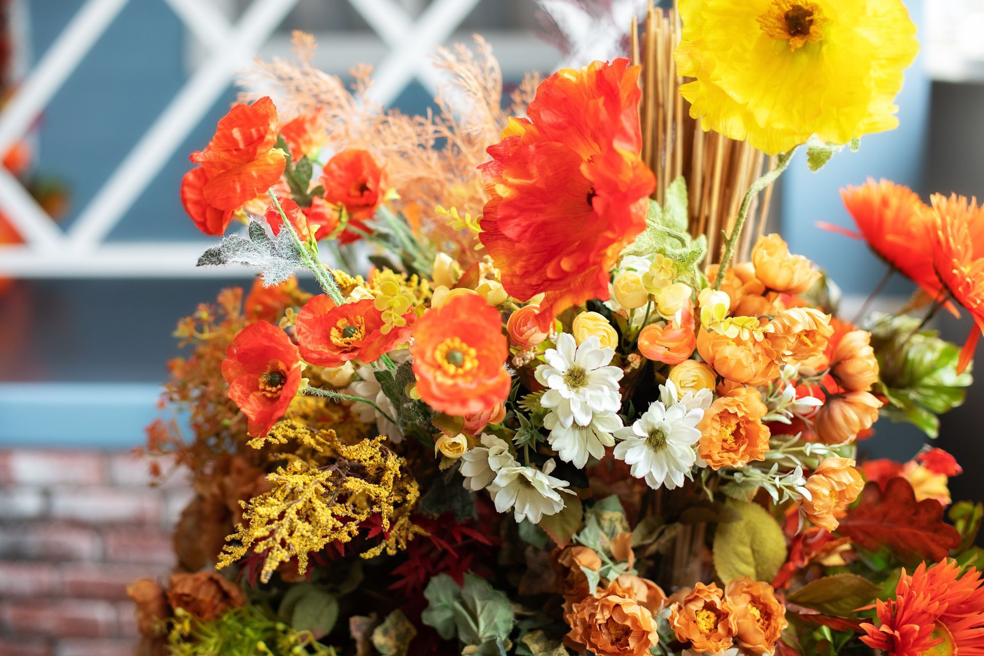 Colorful bouquet of orange, red, white, and yellow flowers in a vase against a brick-and-lattice background
