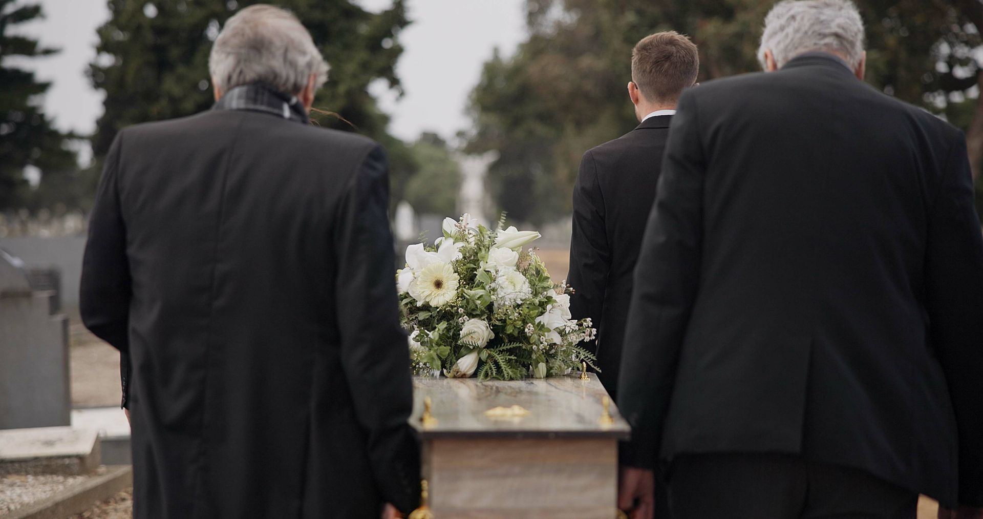 Three mourners in black suits stand at a graveside with a white floral arrangement.