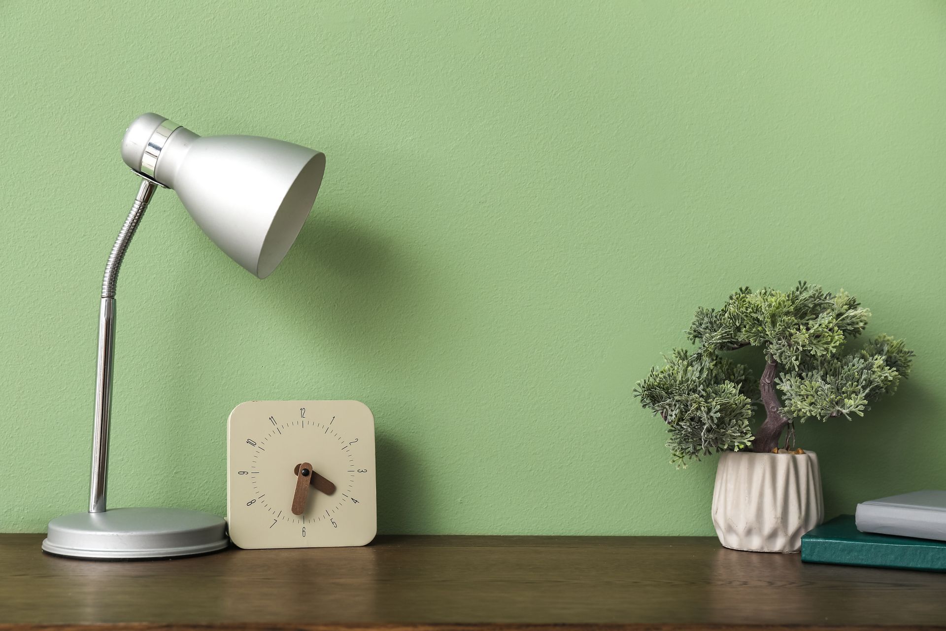 White desk lamp, small clock, and potted plant on a wooden shelf against a green wall.