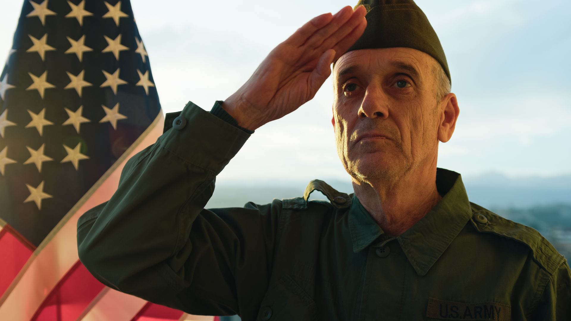 Man in military uniform saluting beside a U.S. flag at sunset