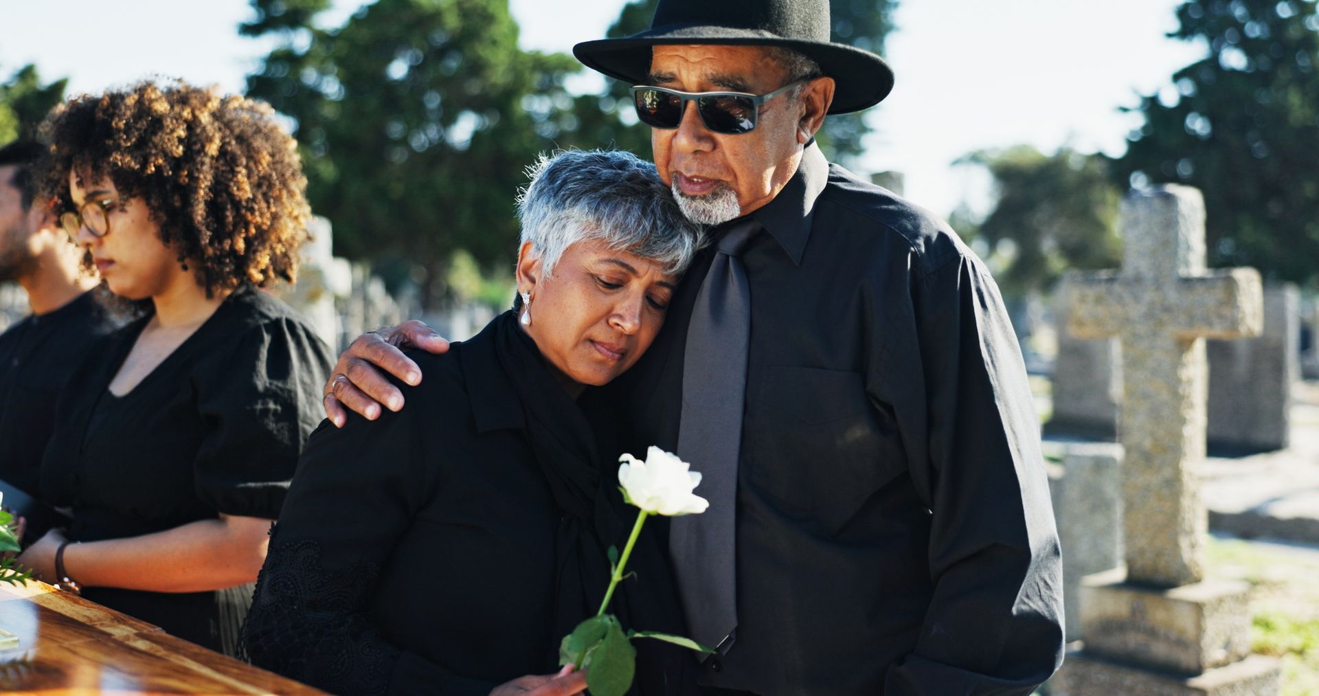 Mourners at a graveside service; one person holds a white flower while another comforts them.