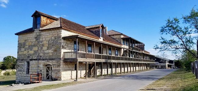 Long, rustic stone building with balconies and a tiled roof beside a paved road under a clear blue sky