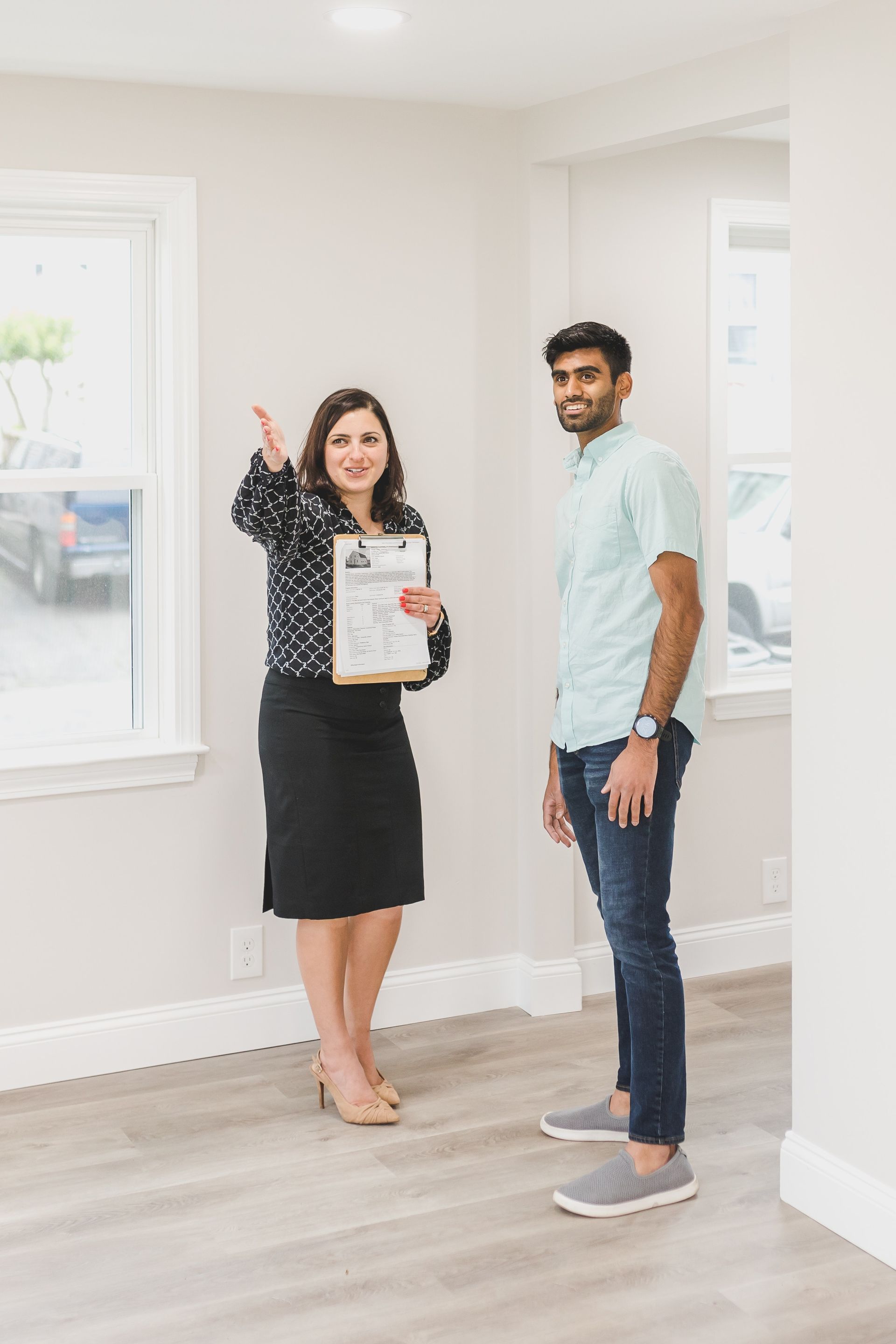 A man and a woman are standing in an empty room . the woman is holding a clipboard and pointing at something.