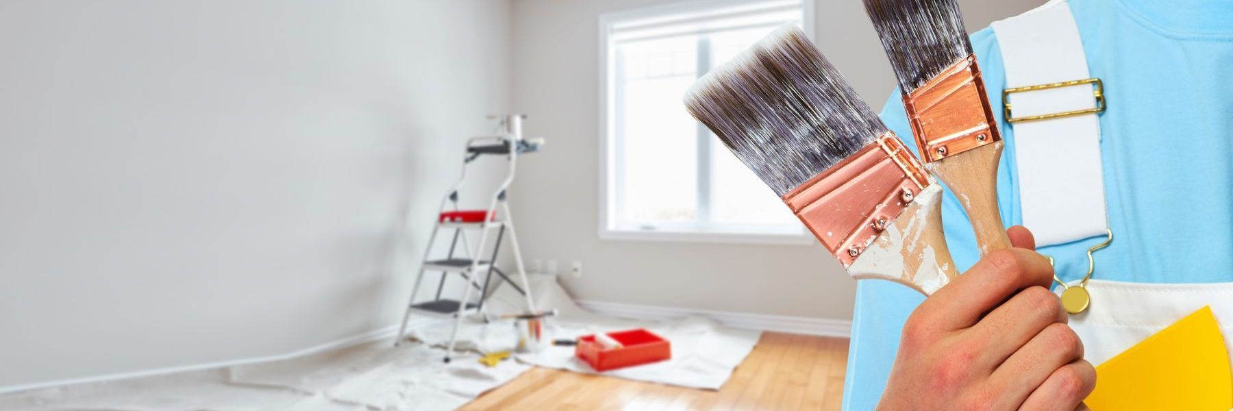 A man is holding two paint brushes in front of a room that is being painted.