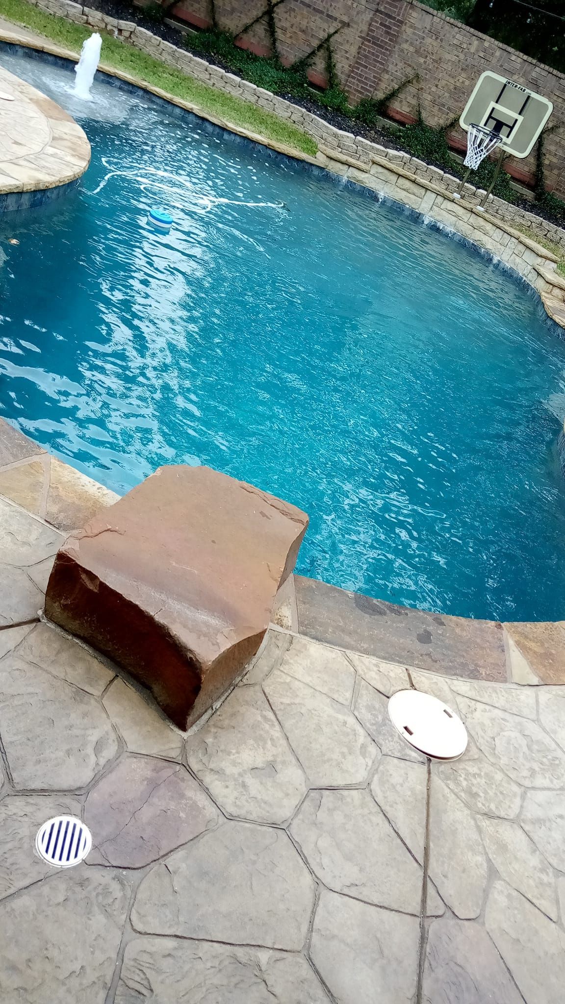 Pool with blue water and stone patio, large rock, and fountain.
