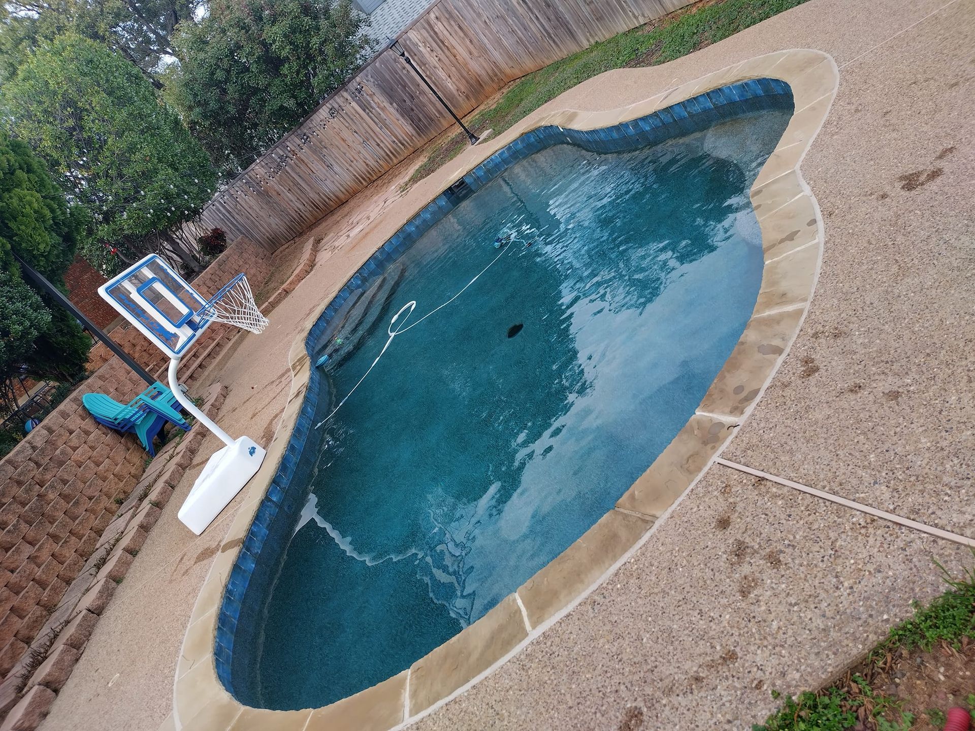 Pool with blue water and tan concrete surround, a basketball hoop, and wooden fence.