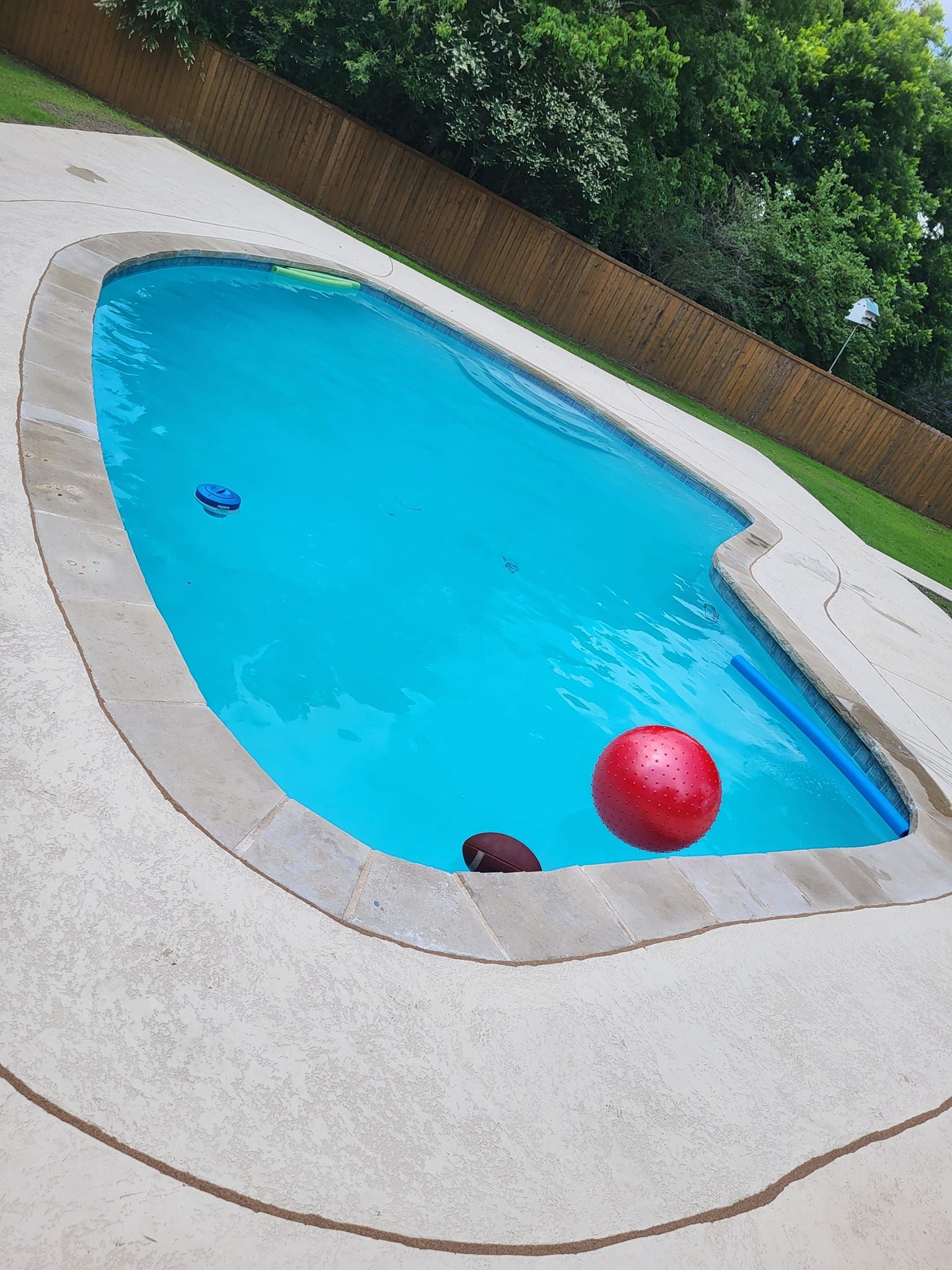 Swimming pool with blue water, red ball, and other pool toys. Concrete border, wooden fence.