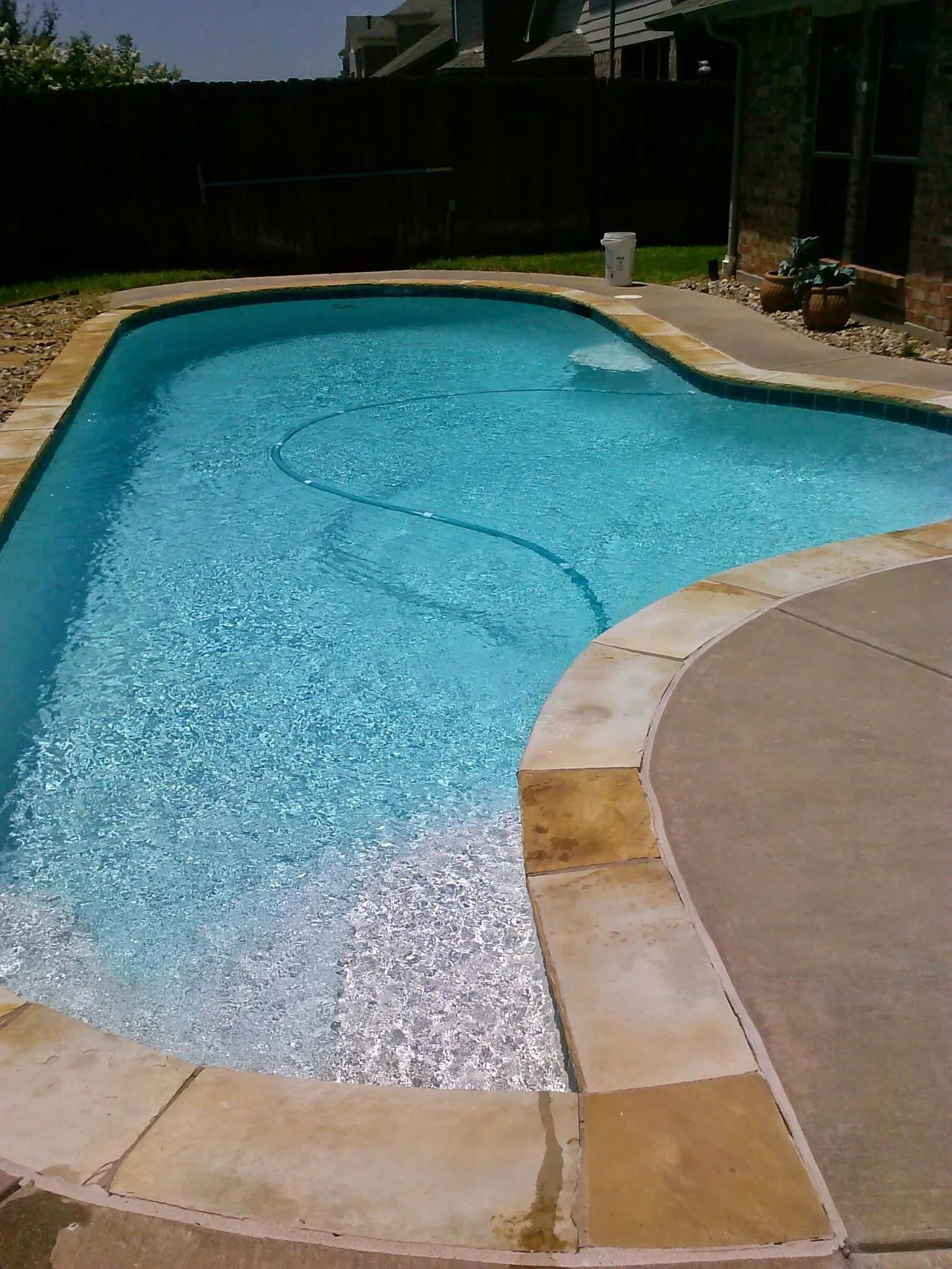 Backyard swimming pool with clear blue water and beige stone decking on a sunny day