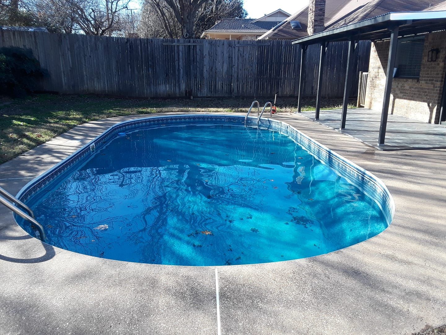 Oval-shaped swimming pool with blue water, concrete surround, wooden fence, and pergola in a backyard setting.