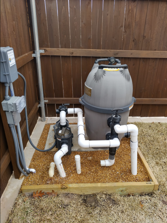 Pool filter system with pump and filter, pipes, and electrical box on gravel base against a wooden fence.