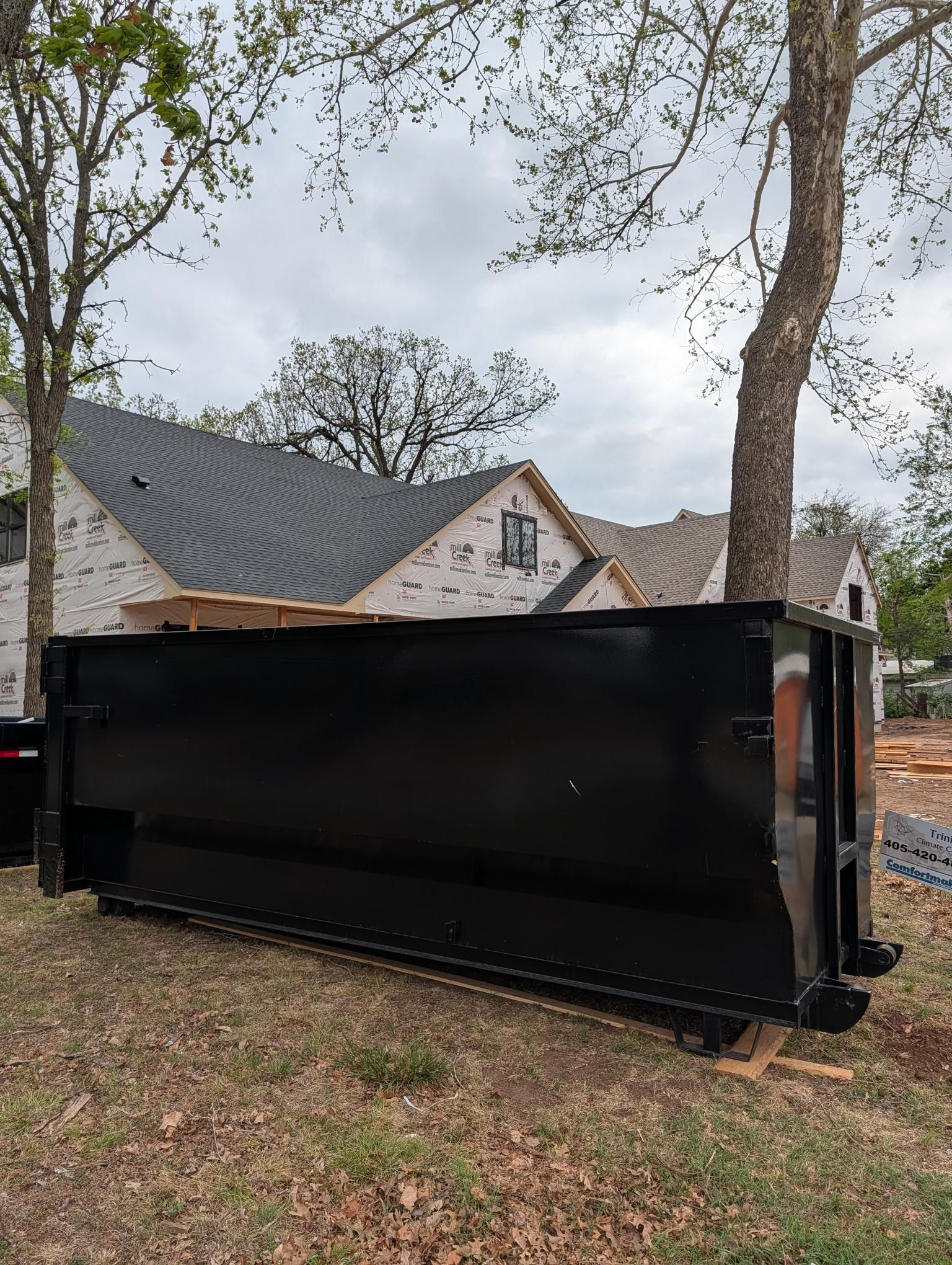 Large black dumpster in a wooded yard beside a house under construction