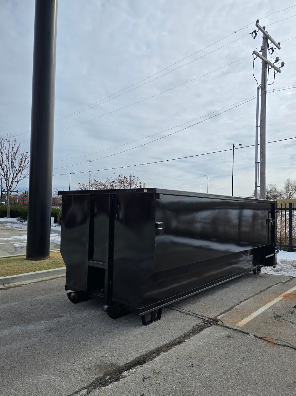 Black dumpster on a roadside beside utility poles and a parking lot under a cloudy sky