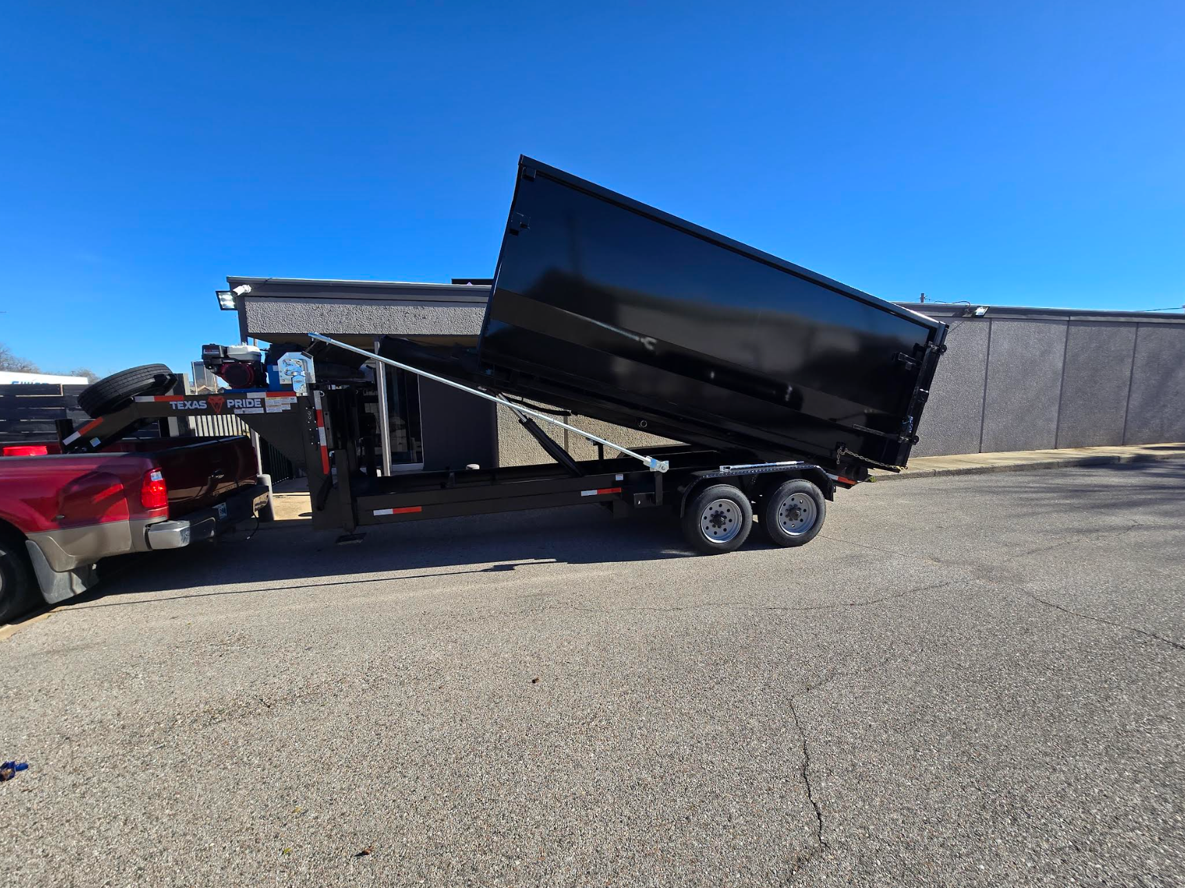Black dump trailer tipped up beside a red pickup in a gravel lot under a clear blue sky