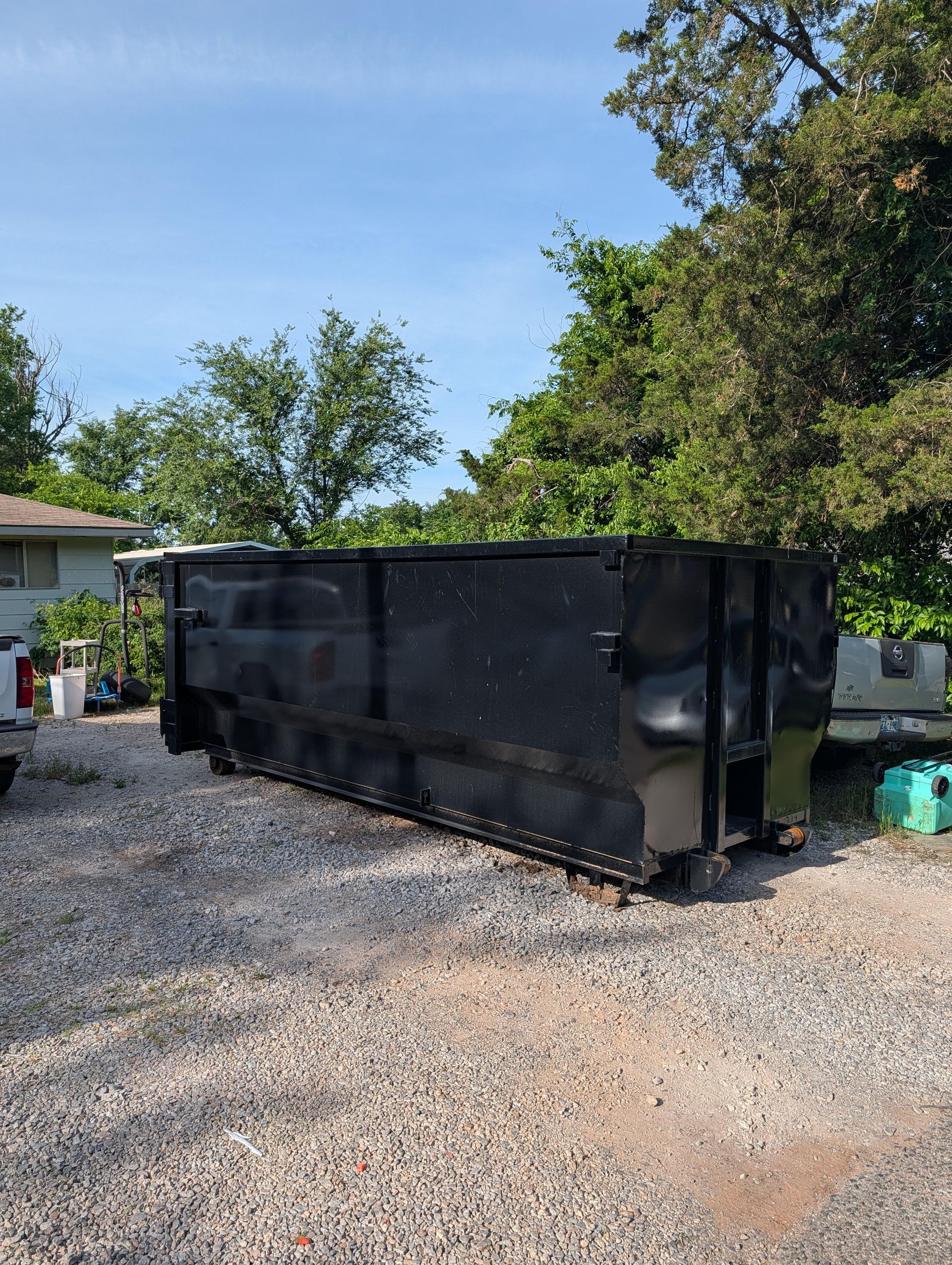 Large black dumpster trailer on a gravel driveway beside a house under a blue sky