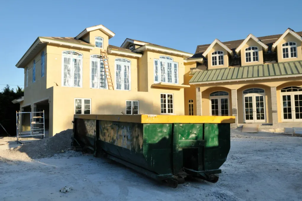 Construction site with a green dumpster in front of a yellow house with multiple windows.