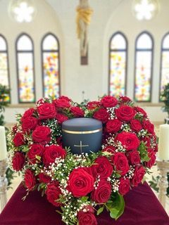 In einer Kirche ist eine schwarze Urne von einem herzförmigen Kranz aus roten Rosen umgeben.