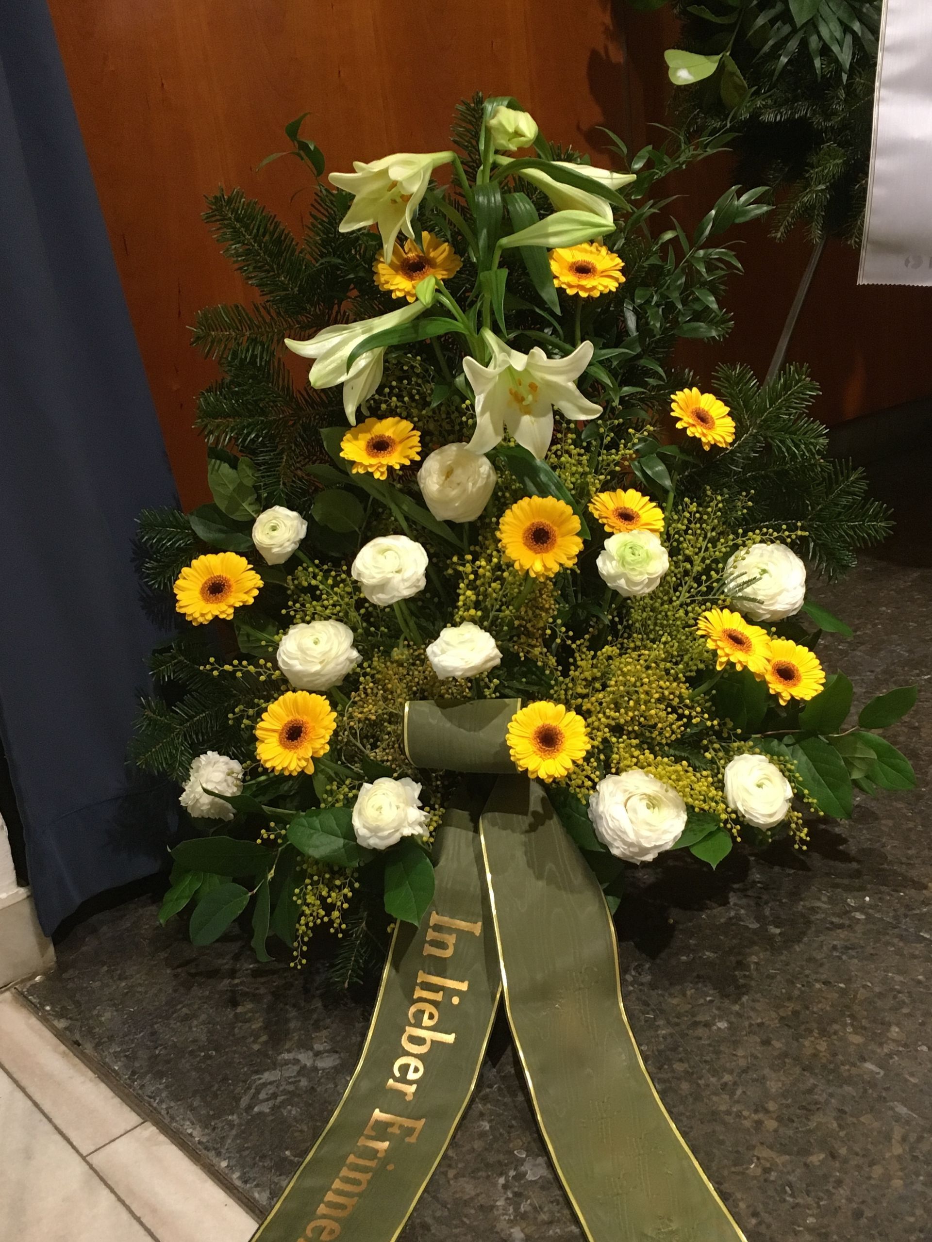 Ein Strauß gelber und weißer Blumen mit einer grünen Schleife mit der Aufschrift „In lieber Erinnerung“