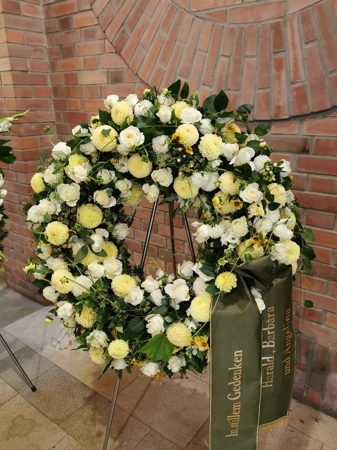 Vor einer Backsteinmauer steht ein Kranz aus gelben und weißen Blumen.