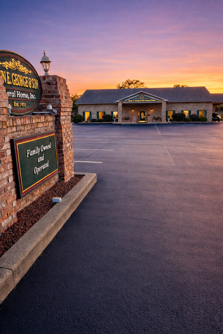 Sunset view of a lodge entrance with a sign and paved driveway under a colorful sky