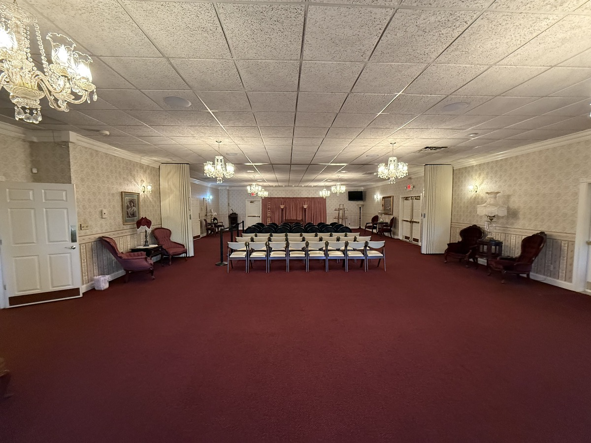Empty banquet hall with burgundy carpet, white walls, chandeliers, and rows of chairs at the far end