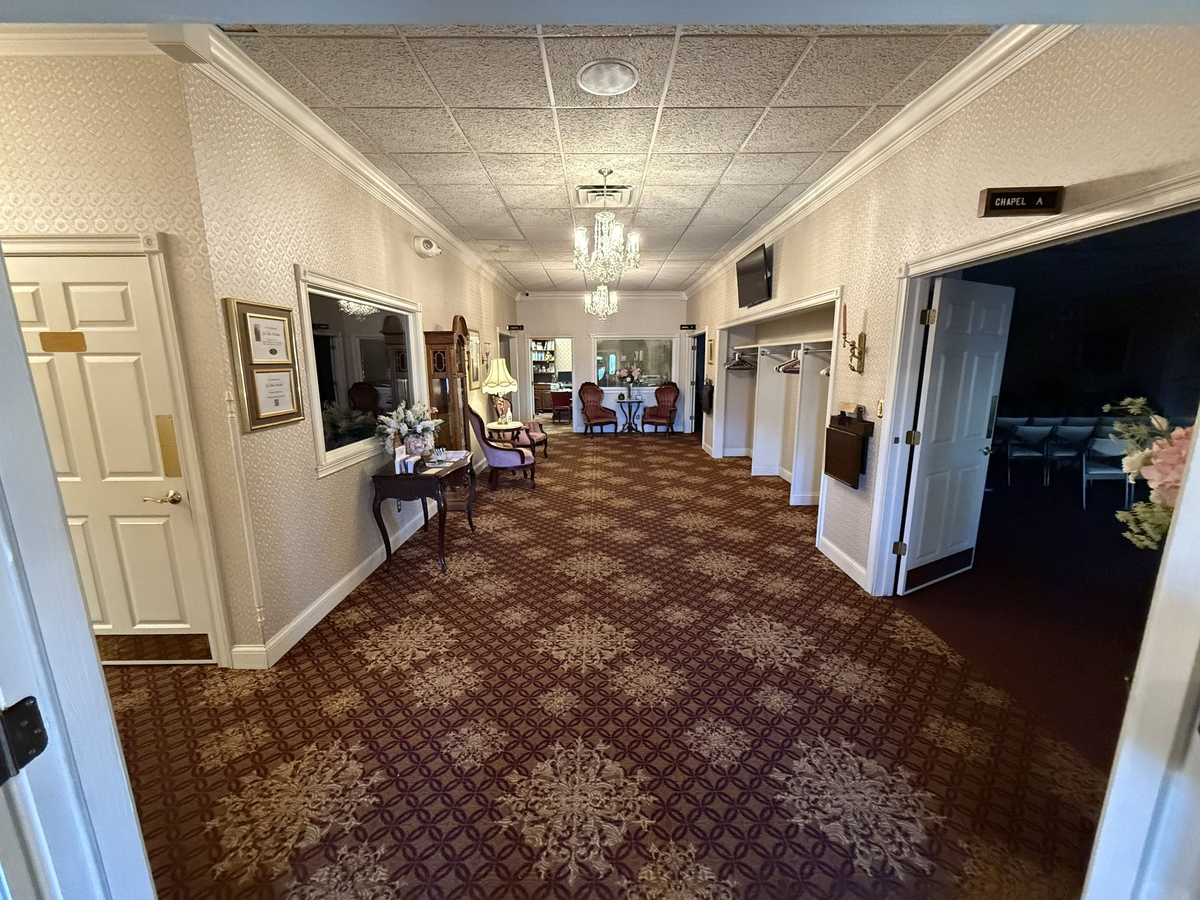 Bright hotel hallway with patterned carpet, cream walls, wall art, and open doorways to rooms
