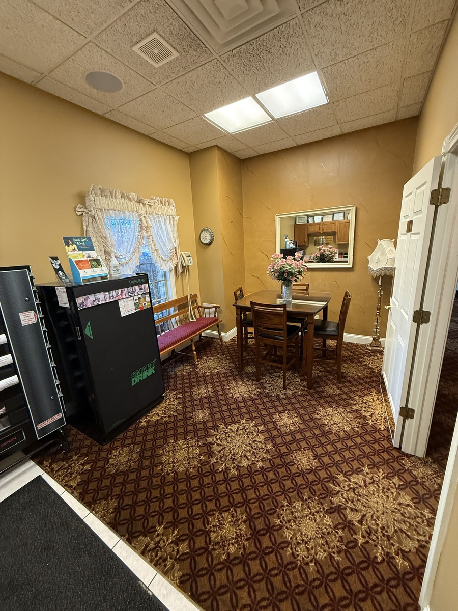 Small waiting room with patterned carpet, black cabinets, table and chairs, white drapes, and warm yellow walls.