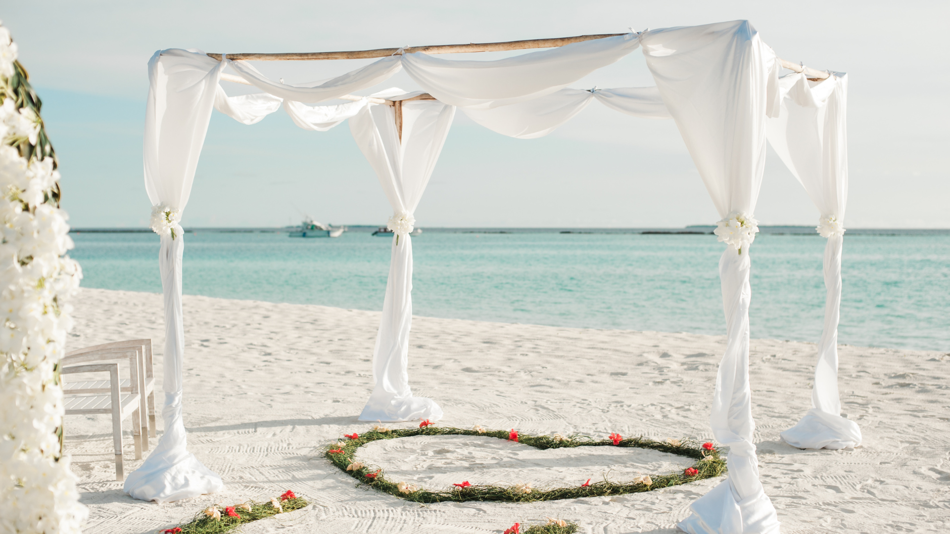 Beach wedding setup: white canopy with draped fabric, heart-shaped floral arrangement, ocean backdrop.
