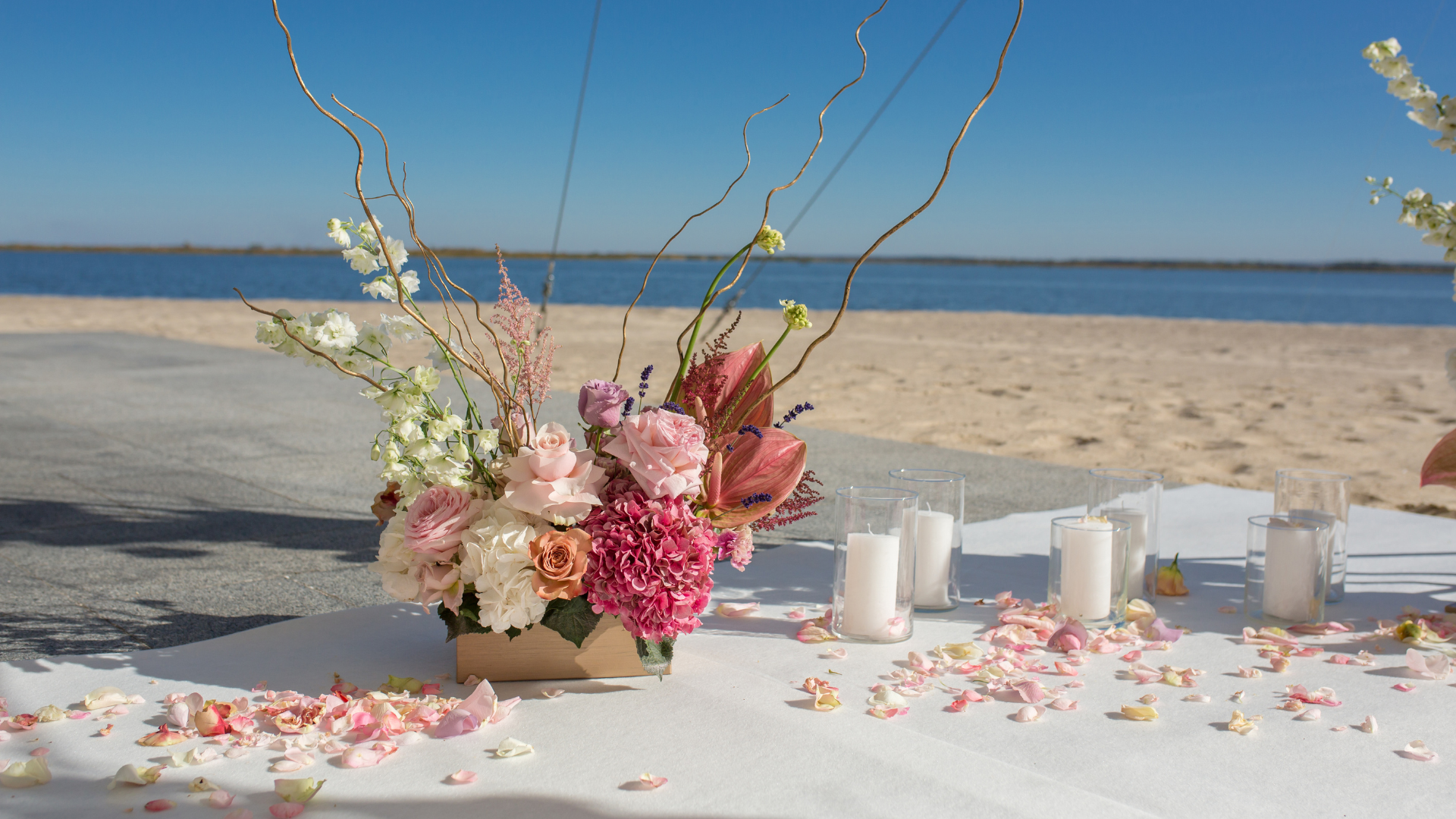 Floral centerpiece and candles on a white table set up on a beach, against a blue sky.
