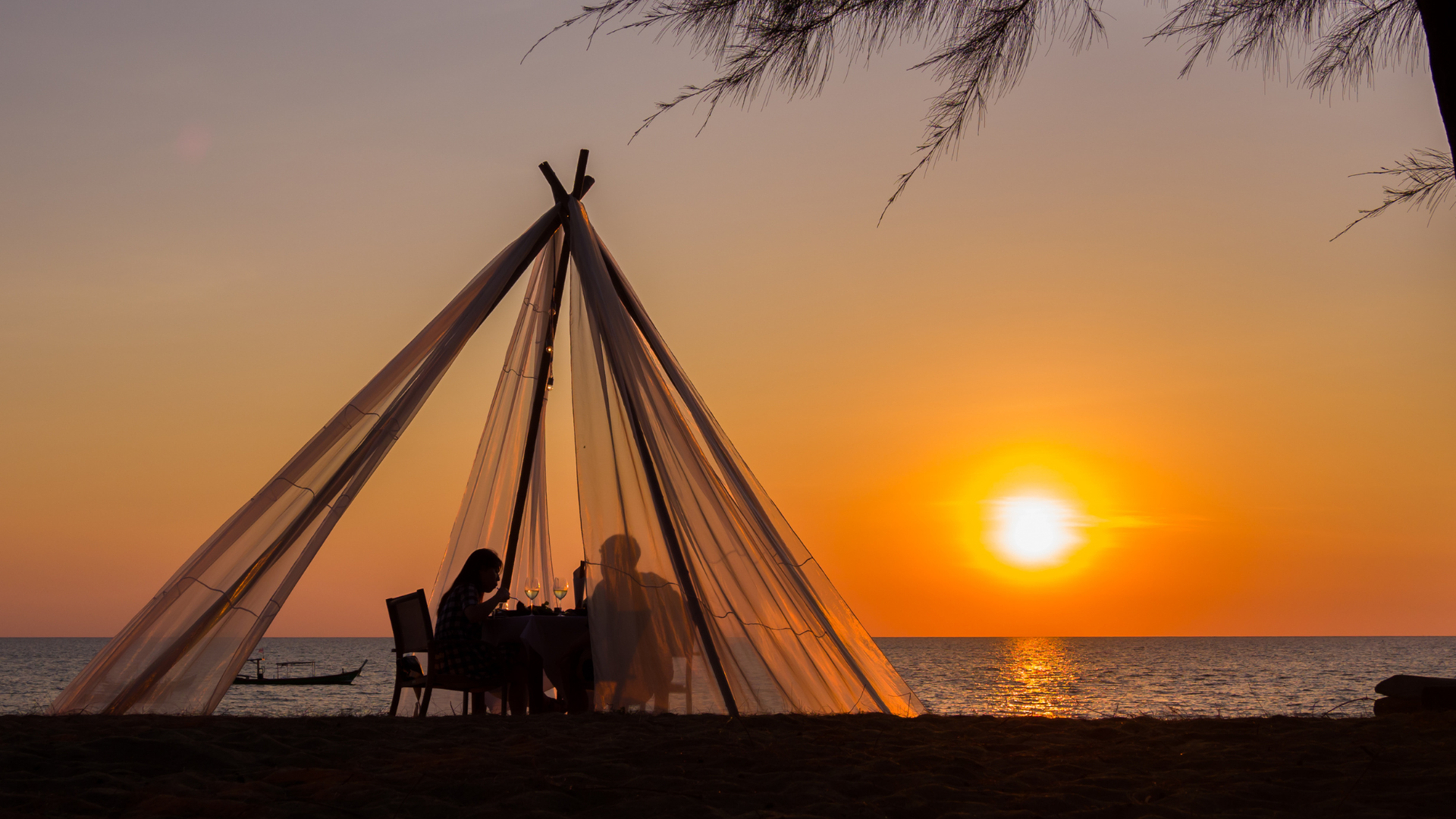 Silhouette of people dining under a draped canopy at sunset on the beach.