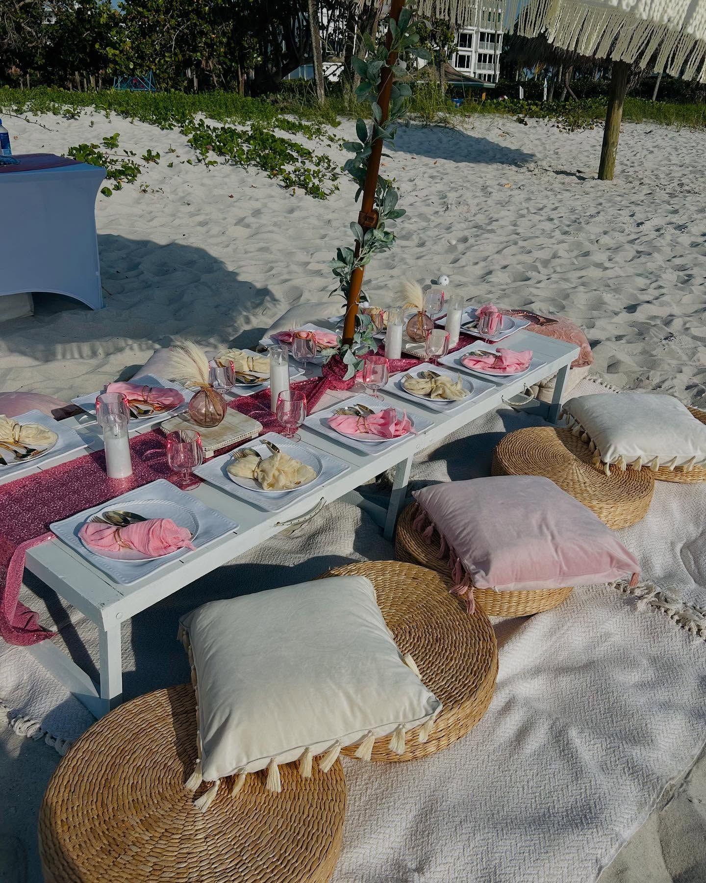 Beach picnic setup with low table, pillows, and food on white sand.
