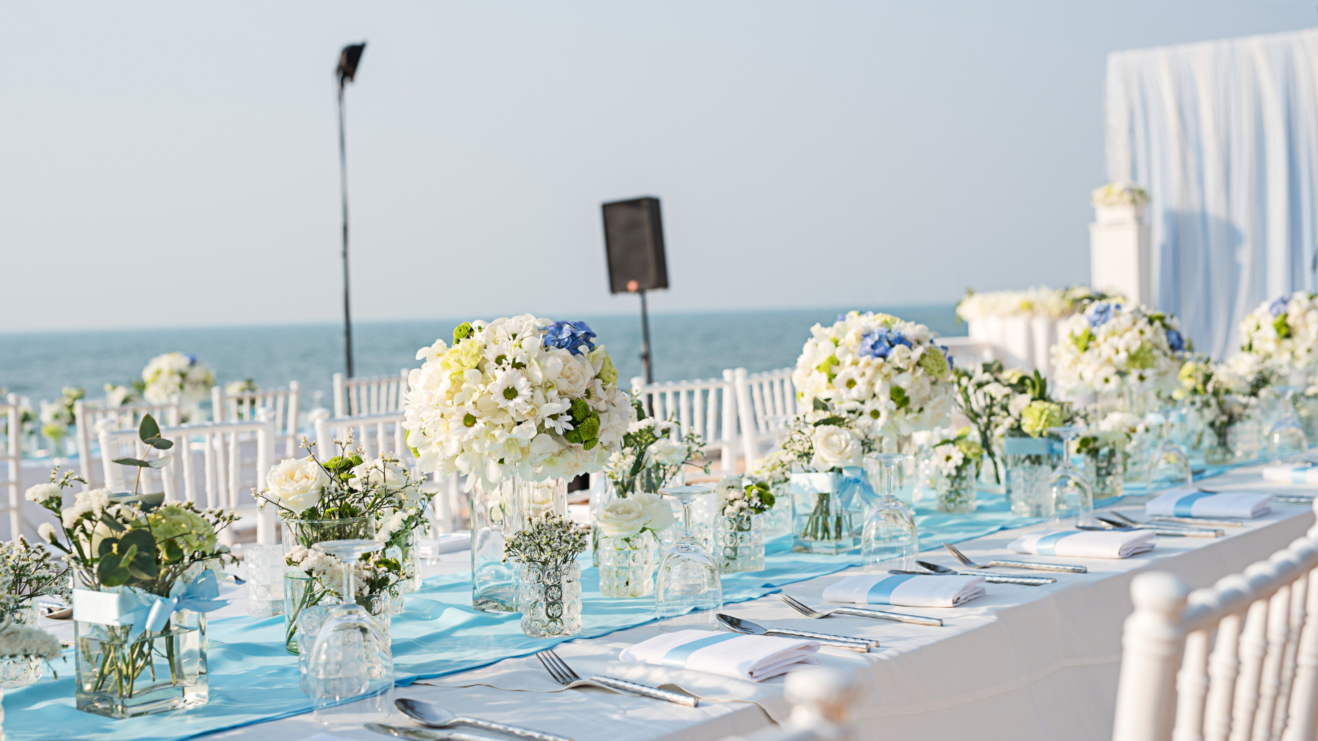 Beach wedding reception table set with white and blue flowers and ocean view.