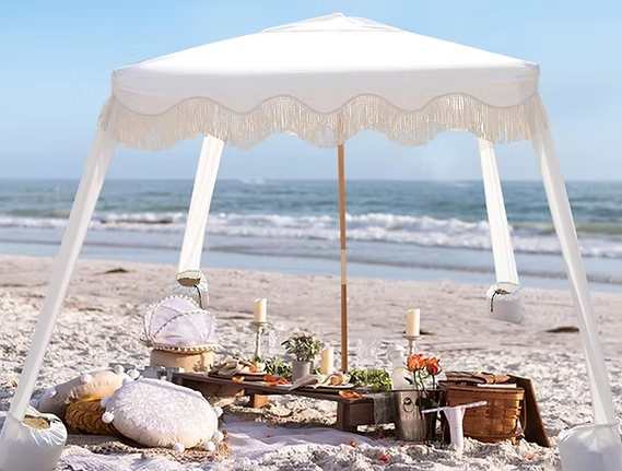 Beach picnic under a white fringed canopy with food, flowers, and candles on a low wooden table; ocean in background.