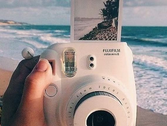 White Fujifilm Instax Mini 9 camera on a beach, printing a photo of the ocean.