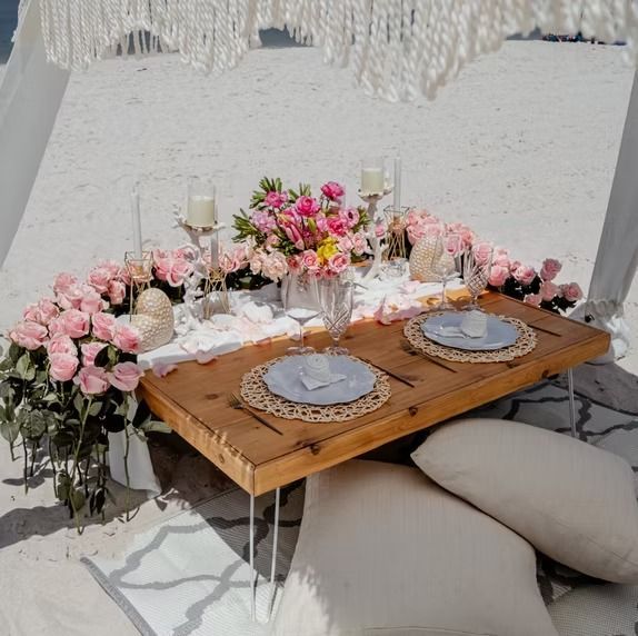 Romantic beach picnic setup with flowers, candles, and a wooden table.