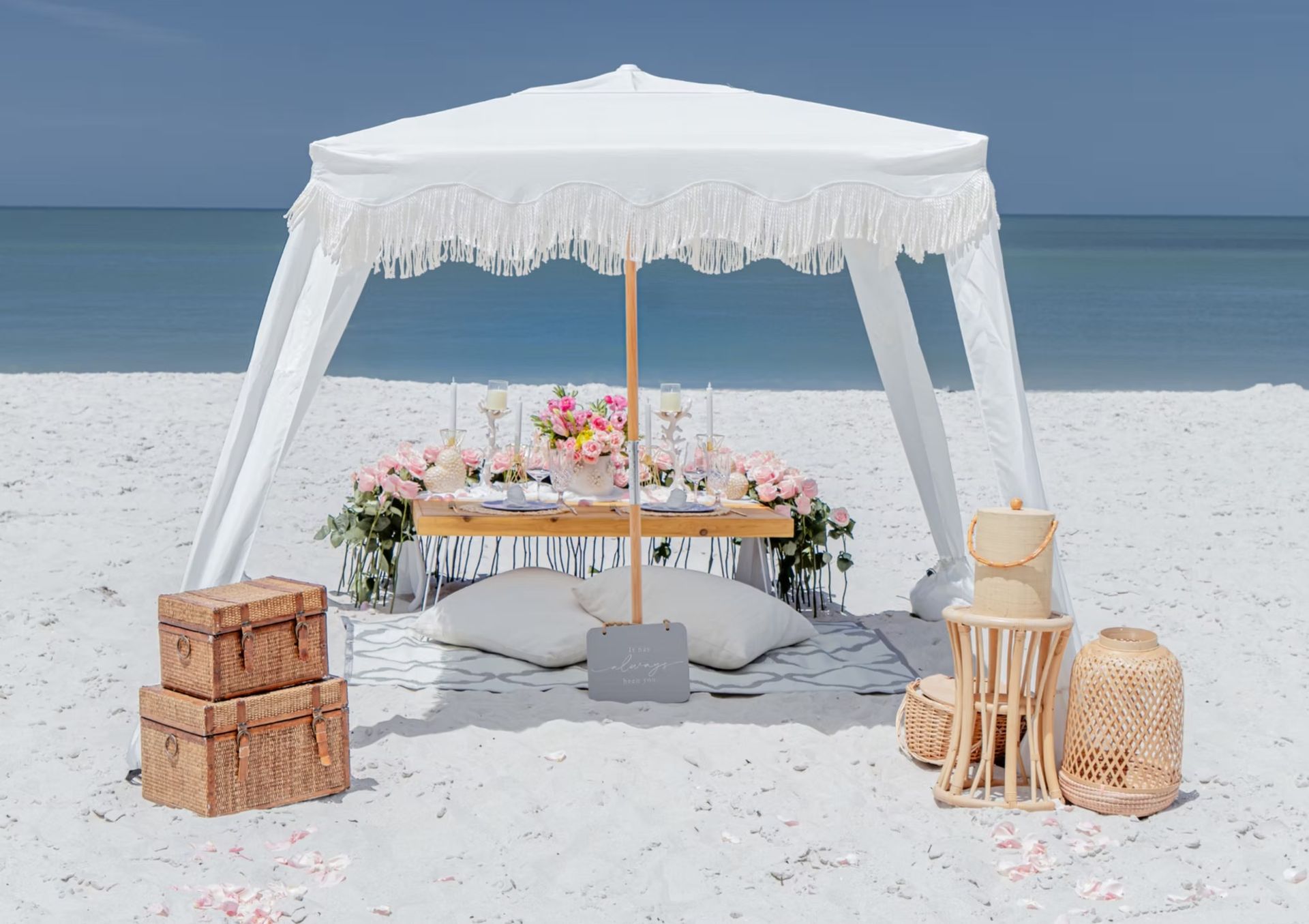 Beach picnic setup with white canopy, table, pillows, and floral arrangements.