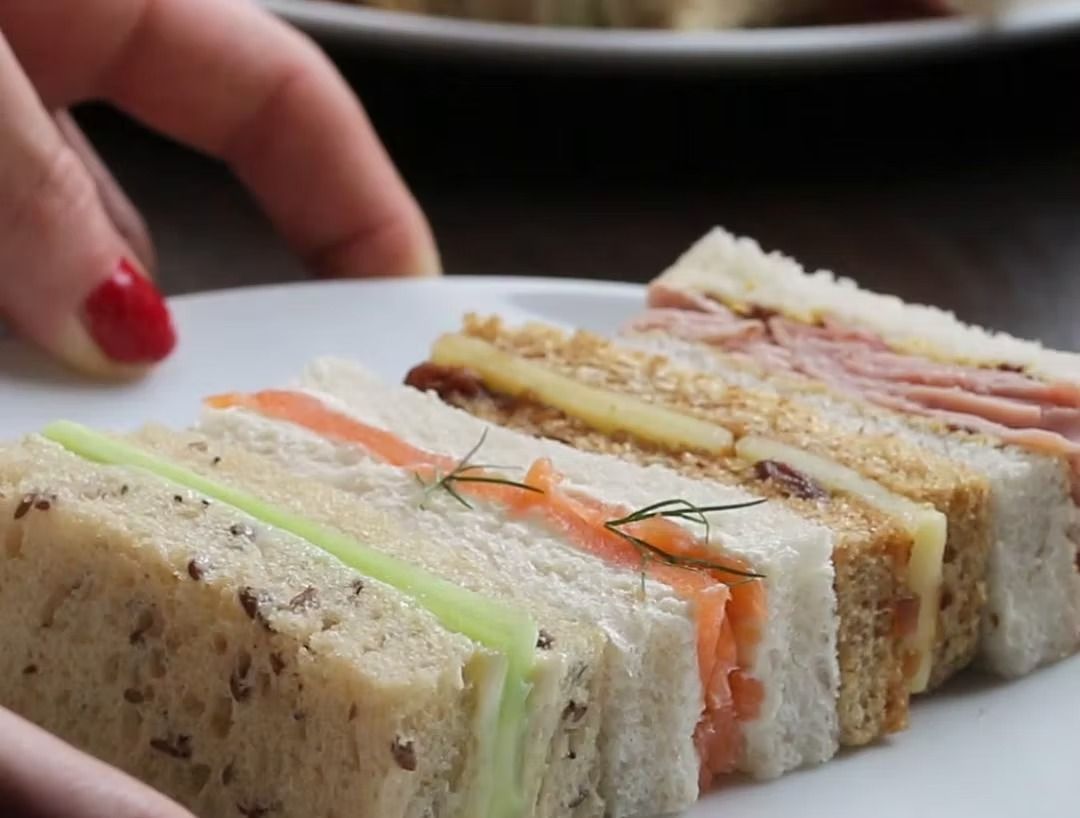 A hand placing a plate with four assorted tea sandwiches: cucumber, salmon, cheese, and ham.