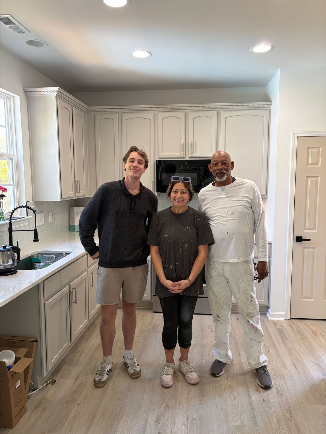 Three people hold up sage green cabinet doors in a workshop, smiling at the camera.